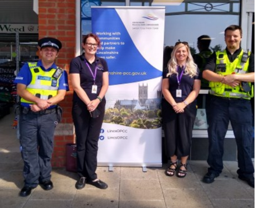 Sunny day. Steph stands in front of a banner displaying Lincoln Cathedral and the PCC's logo, stood alongside Lincolnshire Police Officers.