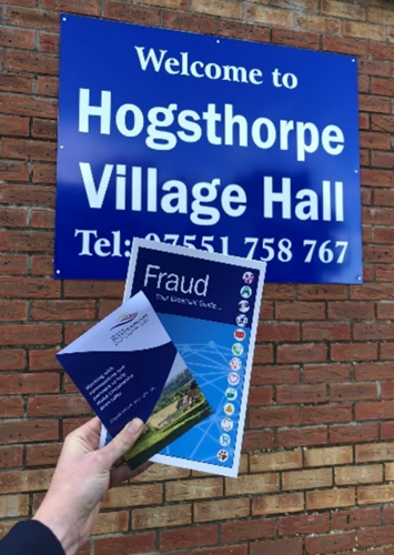 A close up of fraud information leaflets being held against a sign reading: Welcome to Hogsthorpe Village Hall